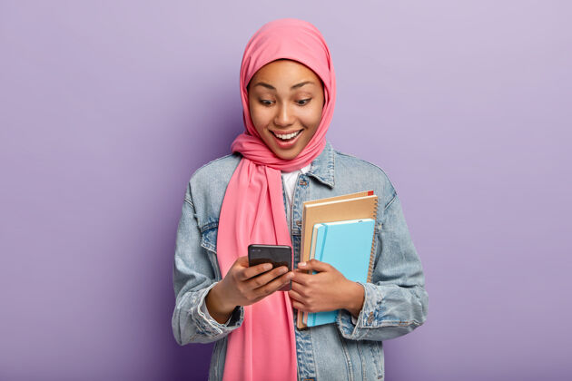 夹克积极的女性看着smatphone设备 戴着粉色头巾 牛仔夹克通讯阿拉伯反应