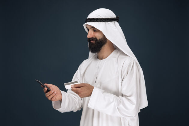 表情阿拉伯沙特酋长的肖像男人用智能手机付账 网上购物或投注信用卡阿拉比人赌博