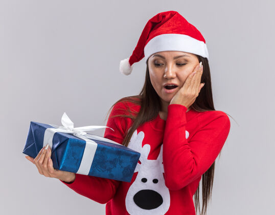 礼物惊讶的年轻亚洲女孩戴着圣诞帽拿着毛衣看着礼盒把手放在脸颊上孤立的白色背景惊讶手拿着