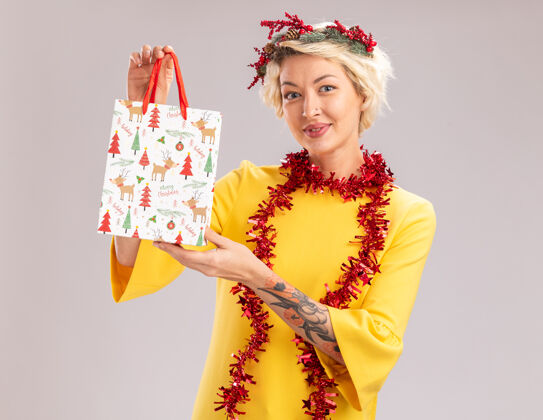 花环年轻的金发女郎戴着圣诞花环 脖子上戴着金属丝花环 手里拿着圣诞礼品袋 看着隔离在白色背景上的相机圣诞节礼物脖子