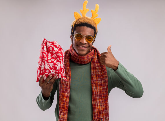 周围快乐的非洲裔美国人 戴着有趣的边 戴着鹿角 脖子上围着围巾 手里拿着红色的圣诞包 手里拿着礼物 微笑着看着相机 竖起大拇指站在绿色的背景上拇指脖子美国