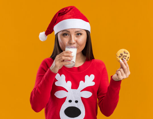 饼干高兴的年轻亚洲女孩戴着圣诞帽 穿着毛衣 拿着饼干 喝着橘黄色背景的牛奶饮料高兴圣诞节