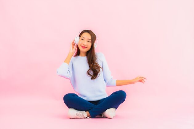 兴奋肖像美丽的亚洲年轻女子用智能手机和耳机在粉红色的墙上听音乐播放列表旋律歌曲