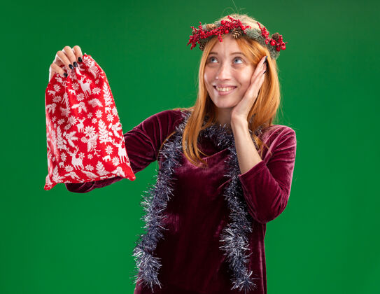 高兴年轻漂亮的女孩戴着红色的花环 脖子上戴着花环 手里拿着圣诞包 手放在脸颊上 在绿色的背景下与世隔绝包脖子抱