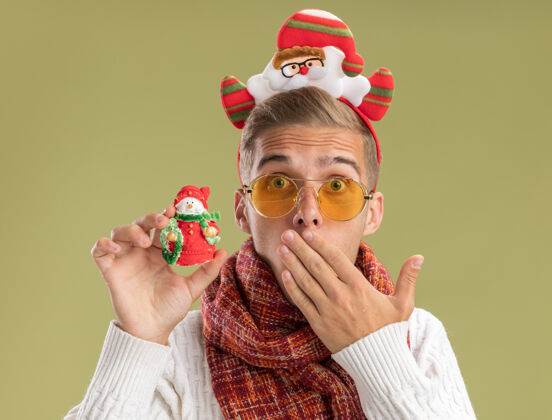 小伙子关心的年轻帅哥戴着圣诞老人的头带和围巾看着相机拿着雪人圣诞饰品手放在嘴上橄榄绿的背景隔离头带圣诞老人看