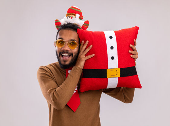 枕头快乐的非洲裔美国人 身穿棕色毛衣 头戴圣诞老人戒指 打着有趣的红色领带 抱着圣诞枕头 站在白色背景下 面带微笑地看着相机脸非洲站着