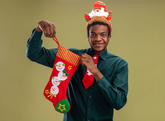 站着一个非洲裔美国人 戴着有趣的圣诞老人戒指 打着红色领带 手里拿着圣诞长袜 站在绿色的背景下 快乐而积极地微笑着看着镜头积极长袜举行