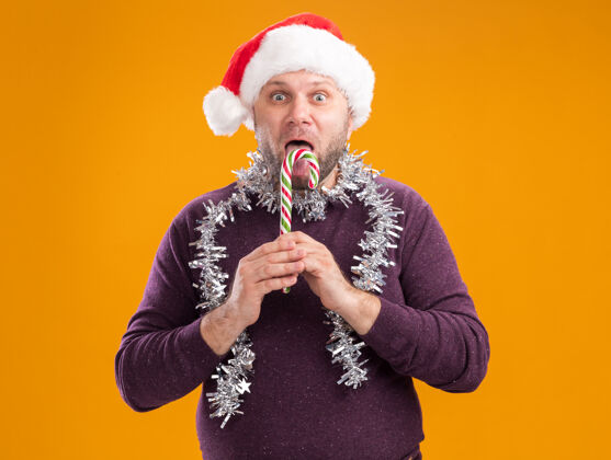 金属片令人印象深刻的中年男子戴着圣诞帽 脖子上戴着金箔花环 手里拿着圣诞甜手杖 看着摄像机 在橙色的背景上显示孤立的舌头圣诞节周围帽子