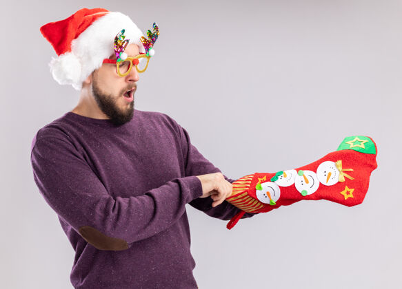 惊讶一个穿着紫色毛衣 戴着圣诞帽 戴着滑稽眼镜 手里拿着圣诞长袜的年轻人站在白色的背景下惊讶地看着它年轻眼镜惊讶