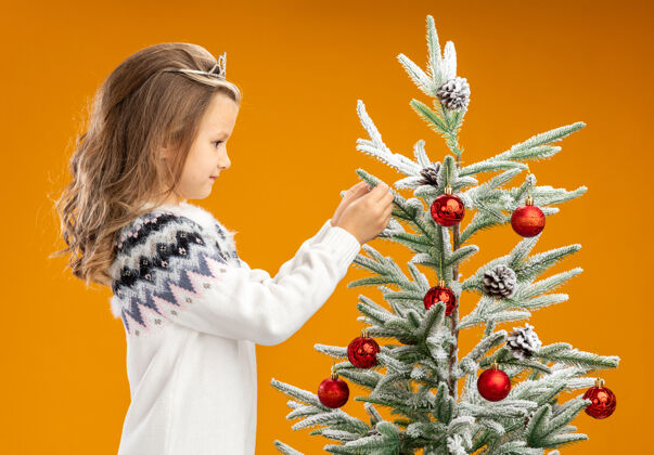 圣诞高兴的小女孩站在圣诞树旁边戴着头饰 脖子上戴着花环 把树隔离在橙色的背景上高兴圣诞快乐圣诞树