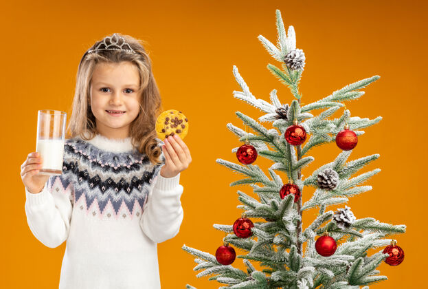 牛奶高兴的小女孩站在圣诞树旁 戴着头饰 脖子上戴着花环 手里拿着一杯牛奶和饼干 背景是橙色的圣诞小快乐