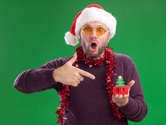 金属片惊讶的中年男子戴着圣诞帽 脖子上戴着金属丝花环 戴着眼镜 拿着并指着圣诞树玩具和日期 看着隔离在绿色背景上的相机日期帽子中年