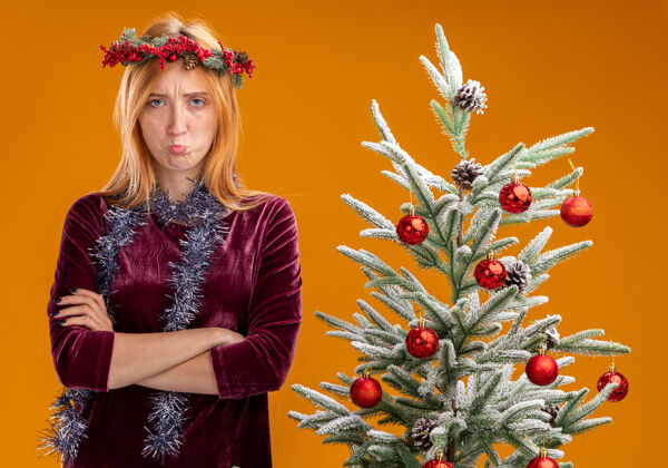 交叉站在圣诞树旁的悲伤的年轻漂亮的女孩穿着红色的裙子 脖子上戴着花环 双手交叉在橙色的背景上年轻树站立