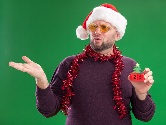 恼火恼怒的中年男子戴着圣诞帽 脖子上戴着金箔花环 戴着眼镜 手里拿着圣诞树玩具 一边看一边 绿色背景上孤立着一只空手眼镜日期圣诞
