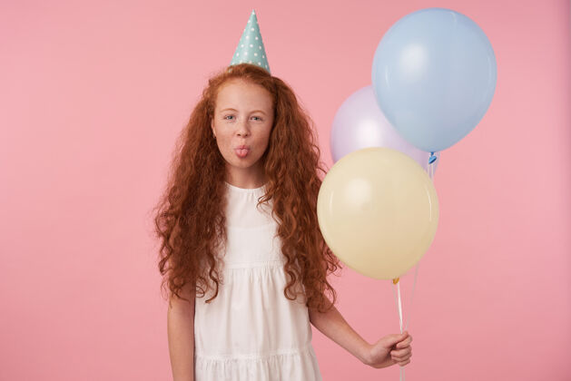 孩子一张室内照片 欢快的红发女孩 长卷发 手持气球站在粉色背景上 身穿白色连衣裙 头戴生日帽 开心地笑着 露出舌头卷曲空气有色