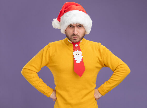 领带戴着圣诞帽 打着领带 手放在腰上 看着紫色背景上孤立的摄像机 躲躲闪闪的白人年轻人帽子年轻圣诞节