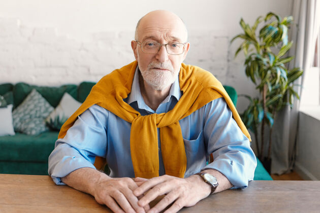 人人 年龄 生活方式和时尚理念帅气的秃头老人戴着长方形眼镜 戴着手表 蓝色衬衫和黄色毛衣坐在木制书桌旁 看着相机看成人室内