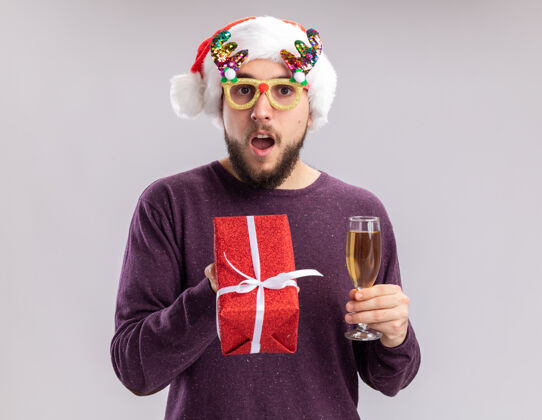 年轻年轻人穿着紫色毛衣 戴着圣诞帽 戴着搞笑的眼镜 手里拿着一杯香槟 惊讶地看着镜头 新年假期的概念站在白色的背景上年站着圣诞老人