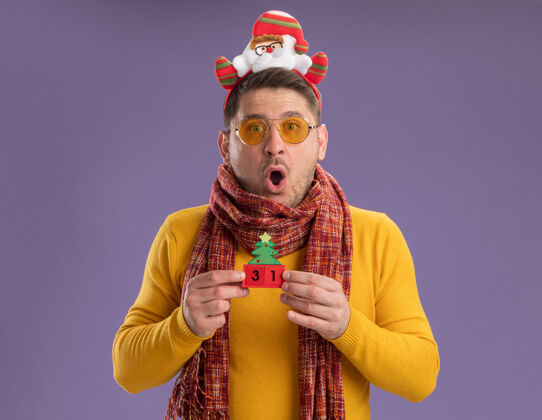 惊讶穿着黄色高领毛衣 戴着暖和的围巾和眼镜的年轻人戴着滑稽的帽沿 头上戴着圣诞老人 他站在紫色的背景上 展示着印有新年日期的玩具立方体温暖年轻眼镜