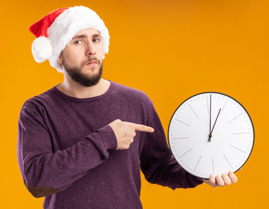 拿着一个穿着紫色毛衣 戴着圣诞帽的年轻人拿着挂钟 用食指指着挂钟 严肃地看着站在橙色背景上的摄像机手指脸帽子
