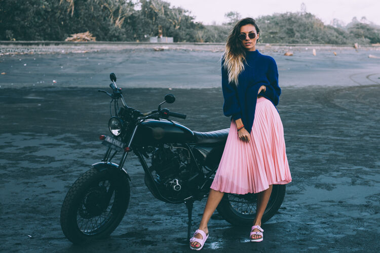 骑戴着太阳镜 穿着蓝色毛衣和粉色裙子的时髦女人的时尚肖像 站在黑色沙滩上的黑色自行车旁骑自行车的女孩交通女孩自行车