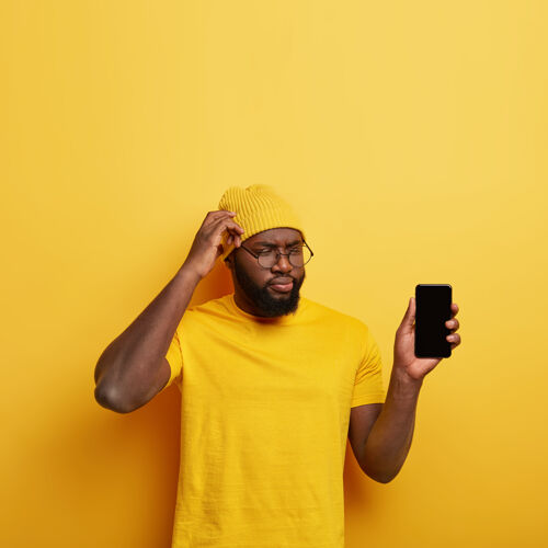 民族迷惑不解的体贴男人搔着头 思考着创建新的应用程序 给你看智能手机屏幕 穿着黄色的衣服沉思垂直剃须