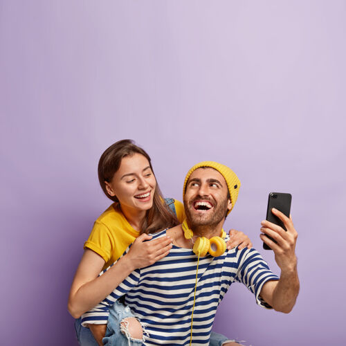 男朋友有趣的年轻夫妇在智能手机上自拍 享受骑在背上的乐趣 有快乐的表情 可爱的女人从背后拥抱男友 在紫色背景下孤立人骑拥抱摄影