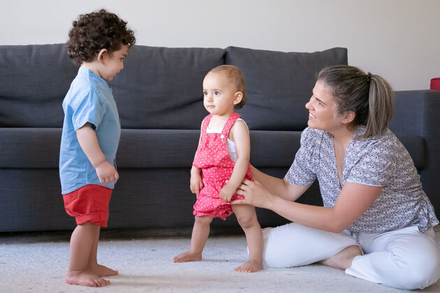 第一快乐的妈妈在家里和孩子们玩 盘腿坐着可爱的小女孩和男孩赤脚站在客厅的地毯上家庭在室内 周末和童年的概念小幼儿可爱