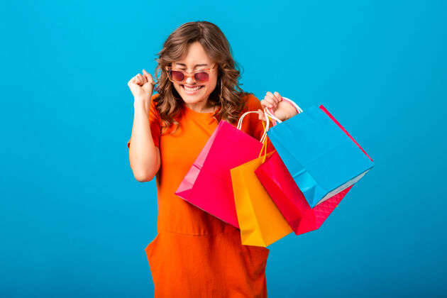 成功图片中的兴奋迷人的微笑时尚女性购物狂穿着橙色的新潮连衣裙拿着购物袋在蓝色的工作室背景隔离女孩女性肖像