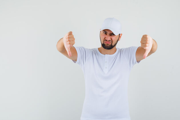 开朗年轻人展示拇指在白色t恤 帽子和看起来不高兴 前视图肖像男性站着