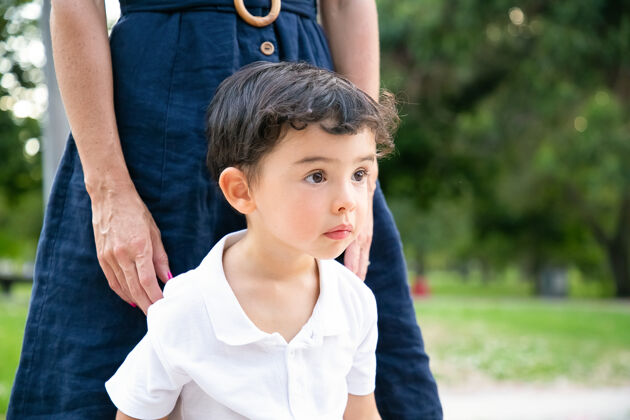 童年专注兴奋的小黑发男孩站在妈妈身边 凝视着户外中镜头童年的概念兴奋肖像孩子