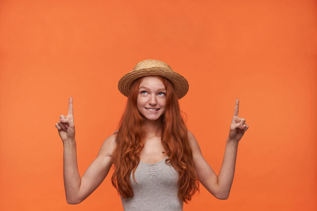 食指摄影棚照片中 身着灰色衬衫 头戴草帽的年轻红发女子站在橙色背景下 抬起食指 咬着屁股向上看发型波浪向上