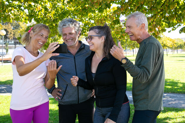 女士快乐女人向健身俱乐部伙伴展示手机屏幕快乐成熟的朋友在公园晨练后站在一起退休或交流的概念肖像手机正面视图