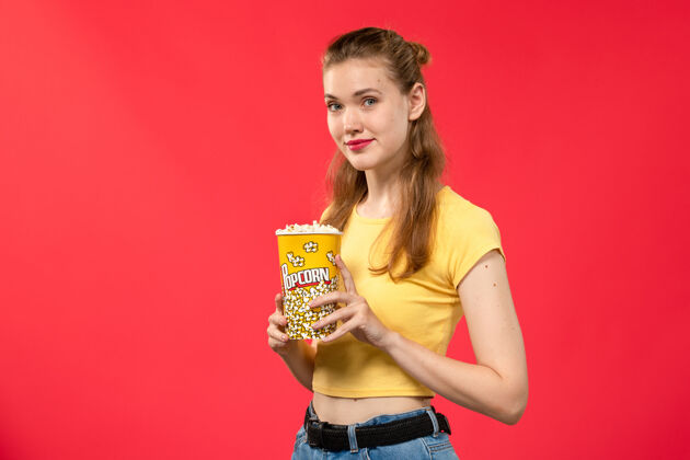 肖像正面图年轻女性在电影院拿着爆米花包 微笑着看红墙电影院的女性趣味电影女性爆米花电影院