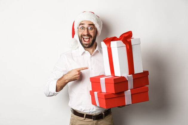 男人圣诞快乐 节日概念惊喜的男人收到圣诞礼物 指着礼物笑着开心 戴着圣诞帽年休闲情绪化