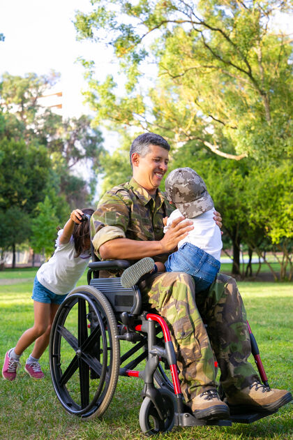 帮助快乐的残疾军人爸爸和两个孩子在公园散步女孩推着轮椅把手 男孩坐在爸爸的膝盖上退伍军人或残疾概念享受把手士兵
