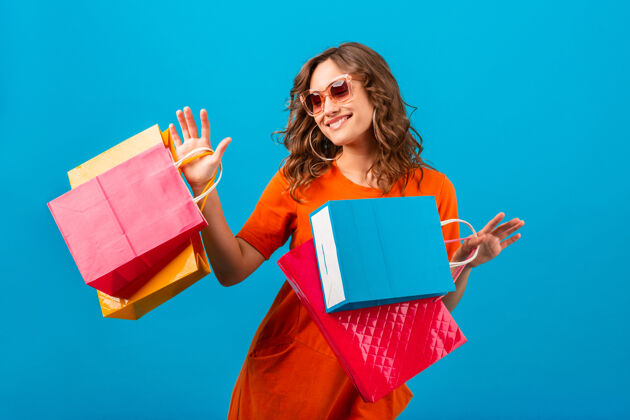 包迷人的快乐情绪微笑时尚的女人购物狂在橙色时尚的超大礼服举行购物袋蓝色工作室背景隔离开朗购物狂购物者