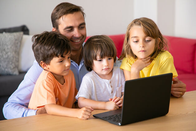 可爱快乐的爸爸和孩子们一起通过笔记本电脑看电影高加索爸爸坐在桌子旁拥抱可爱的孩子男孩和女孩看着屏幕父爱和数字技术概念兄弟玩可爱