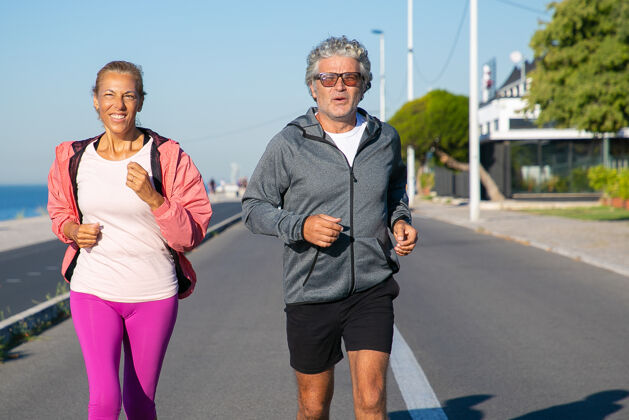 视图开朗成熟的夫妇沿着河岸奔跑白发男女穿着运动服 在户外慢跑积极的生活方式和年龄观念成熟白种人年龄