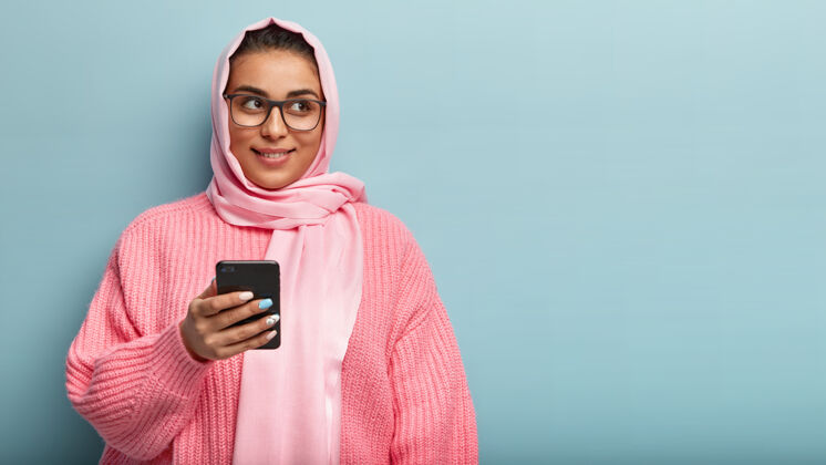 手机在室内拍摄的梦幻积极的年轻女子使用手机 检查信息 穿着粉色针织套头衫 隔离在蓝色墙上的复制空间为您的宣传 阅读文章满意头巾室内