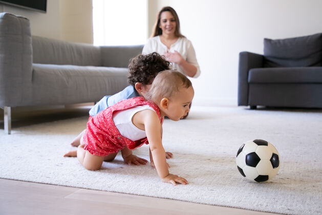 球可爱的小孩爬在地毯上玩足球有爱心的妈妈坐在地板上 微笑着看着孩子有选择性的关注家庭室内 周末和童年的概念婴儿玩妈妈