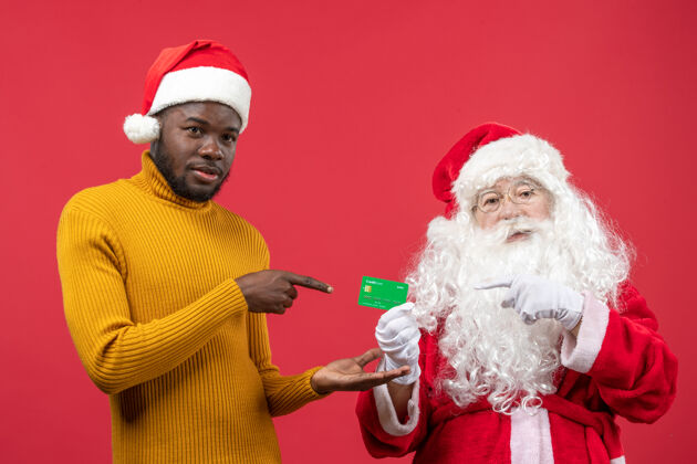 人红墙上的圣诞老人和拿着银行卡的年轻人的正视图前面男人圣诞快乐