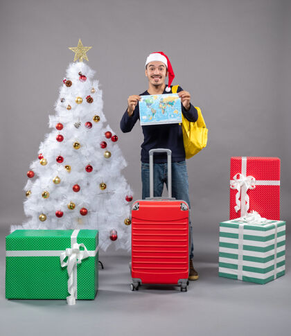 购物者兴奋的年轻人背着黄色的背包 双手拿着地图 靠近灰色的白色圣诞树近圣诞灰色