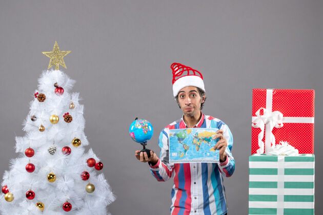 男人前视图年轻人拿着世界地图和地球仪在不同的礼品盒附近圣诞树年轻微笑