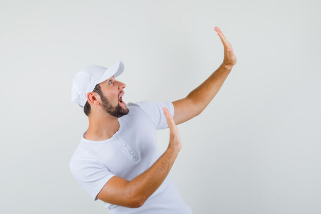 年轻年轻人举起双臂 以预防的方式穿着白色t恤 戴着帽子 看上去很兴奋 前视图人手势工作室
