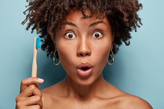 非洲惊讶的黑人妇女的特写肖像用的眼睛盯着 张开嘴 拿着木刷 准备刷牙 惊讶于牙膏的效果 拥有健康的深色皮肤凝视新鲜年轻