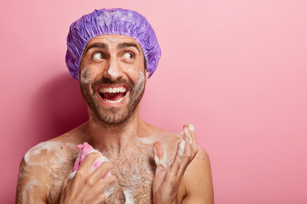 海绵身体护理概念快乐的家伙洗澡 在紧张的工作后放松 用泡沫湿润皮肤 用海绵擦胸脯 愉快地看着旁边 隔离在粉红色的墙上满意男性性感