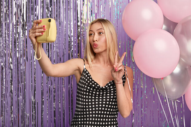 折叠快乐的金发女人化妆 嘴唇合拢 用小相机自拍 站在装饰着气球和金属丝的墙上 穿着时髦的裙子 展示和平的手势化妆风格年轻