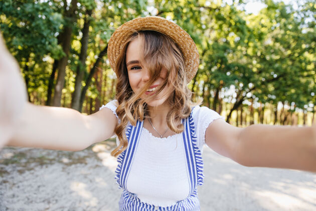 金发欧洲可爱的卷发女孩在公园里嬉戏迷人的戴帽子的女士在树上自拍欧洲卷曲夏天