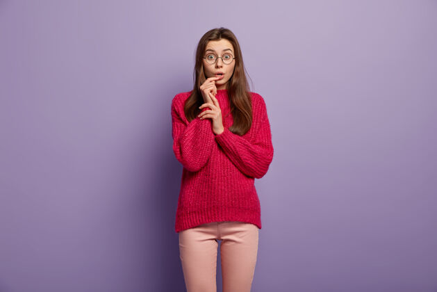 眼镜印象深刻的少女紧握着张开的嘴 惊愕地盯着 戴着圆眼镜 红色的套头衫 孤立在紫色墙壁上的人 反应 表情裤子青少年惊奇
